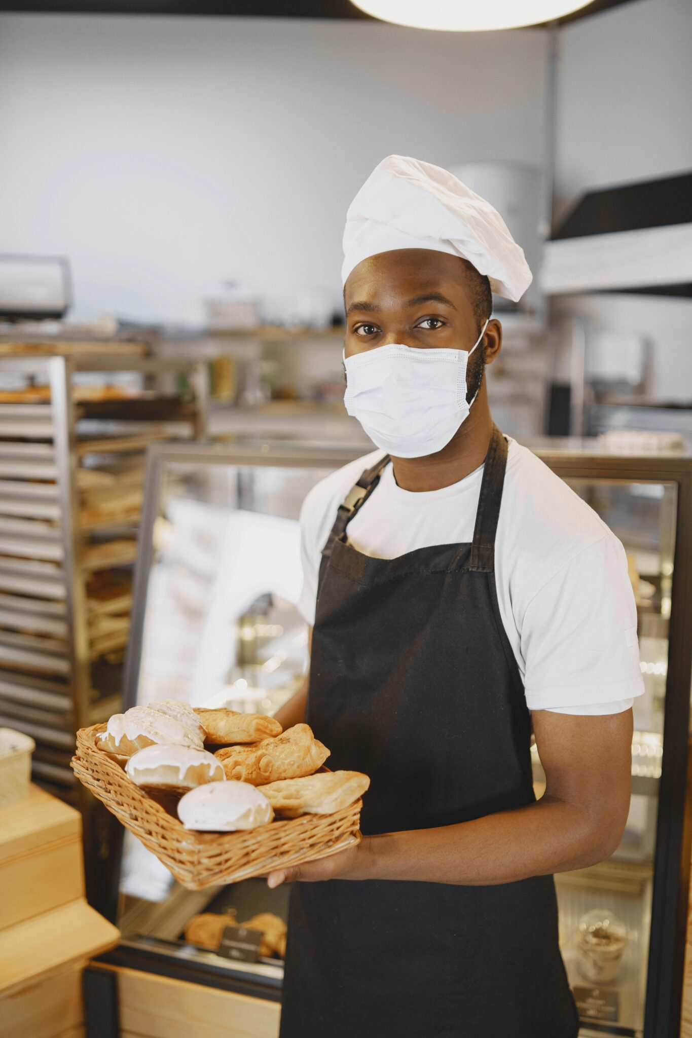 Baker with face mask displaying fresh pastries in a modern bakery.